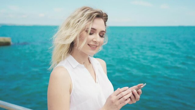Young attractive blonde girl in a white blouse and blue jeans, smiling, looking at the camera and flipping through a photo on the phone on a background of the sea.