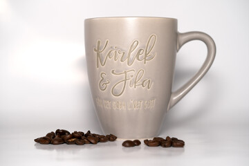 Coffee cup says "Love and Fika" on a white background with coffee beans infront