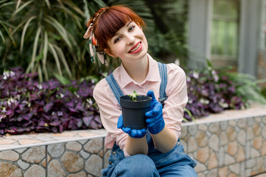 Portrait of smiling red haired girl greenhouse worker, wearing gloves holding little black pot with succulent seedling in hands, and posing to camera on the background of tropic plants