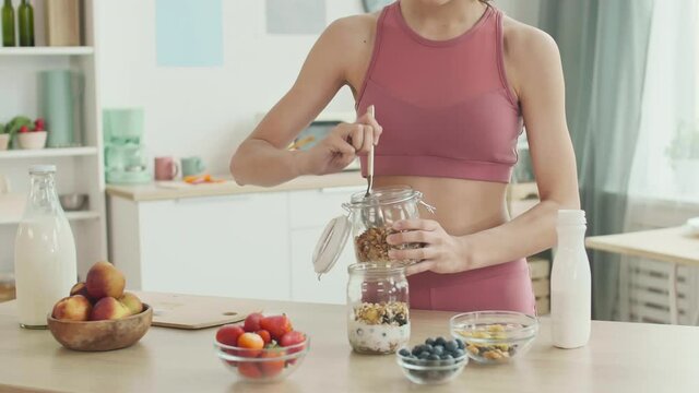 Medium shot of unrecognizable sportswoman standing at kitchen table and making granola with yogurt and fresh berries for breakfast