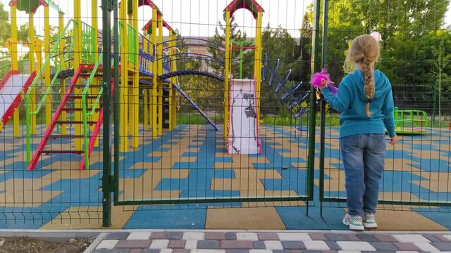 Sad lonely child near the closed playground because of the quarantine.A girl with long blonde braided hair staying alone near empty fenced showground without children.COVID-19 lockdown social distance