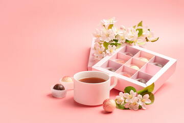 Chocolates of various colors in a festive elegant box packing a cup of tea on pastel pink background decoration flowers.