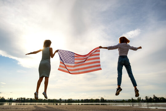 Two young friends women holding USA national flag jumping up together outdoors at sunset. Silhouette of girls celebrating United States independence day.
