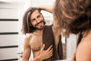 Handsome young man with a smile looks at himself in the mirror in the bathroom after a shower....