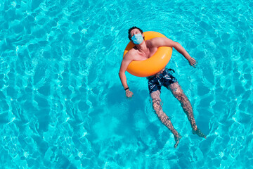 Caucasian young man is floating / swimming in the pool with a protective face mask. The concept of Personal protective equipment during the Covid-19 pandemic or coronavirus infection. Top view.