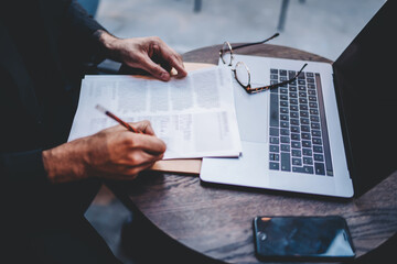 Cropped image of man’s hands noting ideas on paper sitting at wooden table in coffee shop, male...