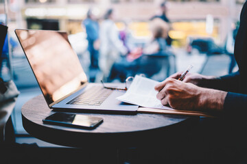 Cropped image of man’s hands planning strategy making notes on paper working remotely in cafe...