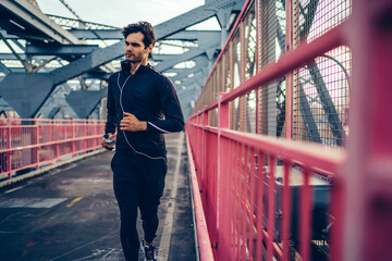 Sporty man with muscular body running across city bridge with bottle in hands, young jogger enjoying cardio exercises and workout outdoors, european athlete making effort for achievement of goal