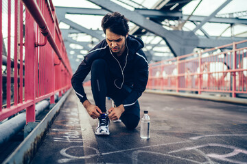 Fototapeta premium Runner man getting ready to run tying sneakers laces on bridge while listening audio records via electronic headphones, european jogger in sport tracksuit preparing for cardio exercises outdoors