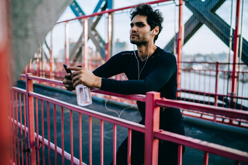 Fototapeta premium Thougtful young athletic man in earphones listening motivated audio songs via telephone player after workout in urban setting.Professional sportsman in active wear standing on bridge during break