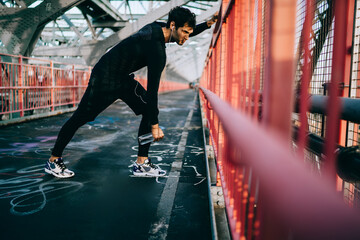 European sportsman in trendy tracksuit stretching on city bridge before start running across street...