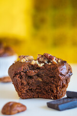 Healthy muffin with a bite from the center a homemade freshly baked and gluten free with pecans and cocoa powder and chocolate cubes on white table with yellow blurred background, nutrition concept