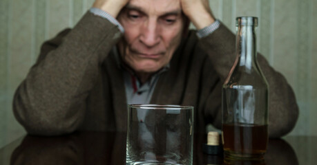 sad senior man sits near bottle with whiskey and empty glass holding hands on head