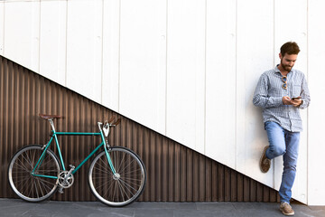 Handsome young man with mobile phone and fixed gear bicycle standing on a two-color modern wall.