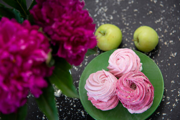 Pink marshmallows in a green plate on a background of green apples and lilac peonies.