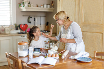 Grandma in white apron with cute curly granddaughter spend wonderful time in the kitchen, smiling and there are happy.