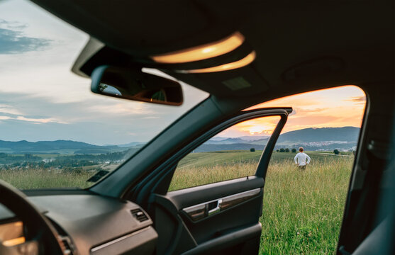Young man standing in high grass and enjoying the sunset sky colors through the open car dor view. Traveling by car concept image.