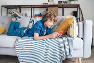 Preteen boy lying at home on cozy sofa dressed casual jeans and shirt. listening to music and chatting using wireless headphones connected with smartphone. Child using electronic devices concept © Soloviova Liudmyla