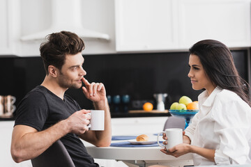 Beautiful woman holding cup of coffee and looking at handsome boyfriend in kitchen