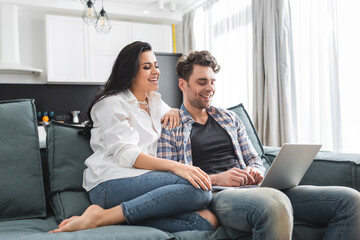Smiling woman sitting near handsome boyfriend using laptop on couch at home