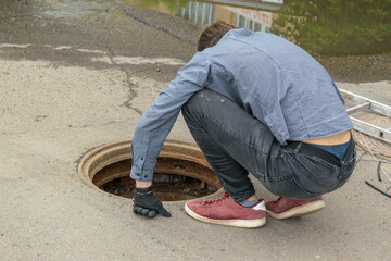 Man carefully looks at  open Manhole. Squatting bent over. View from the back. Concept of...
