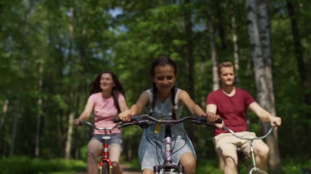 Slow motion panning shot of happy little girl enjoying cycling with parents in forest. Active daughter riding bicycle faster than mother and father
