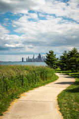 Fototapeta na wymiar A curved path with vegetation and trees on either side leading towards a view of the Chicago skyline with highrise buildings and architecture with blue cloudy sky above.