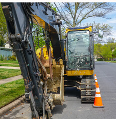 Front view of a yellow heavy equipment excavator parked on the side of a residential street in front of a house with orange warning cones in front