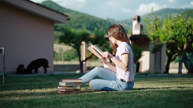 Pensive female student sitting on the grass near the house among the books and reading story. Concentrated girl reading book, turning pages and analyzing information. Preparation for exam outdoors