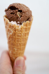 Hand holding a waffle cone with a scoop of chocolate ice cream on top of it, white background.