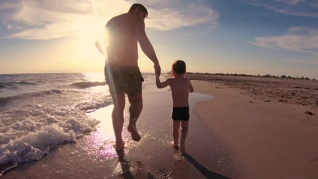 happy father and toddler baby boy running along the sandy beach in the waves on the sunset