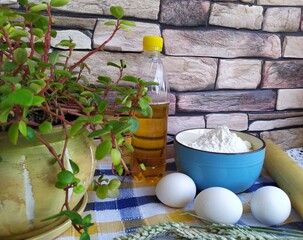 Still life of food in the kitchen eggs a bowl of white flour cookies on a plate a wooden rolling pin grains ears of wheat a bottle of sunflower oil a room flower near the window