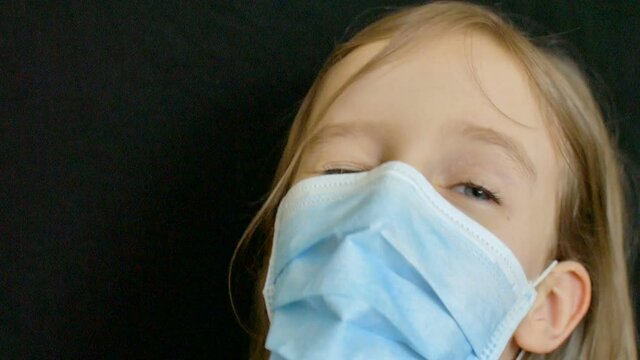 Little blond girl with blue eyes and long hair is posing on black backgound in surgical disposable mask for protection of Coronavirus Covid-19 during self-isolation while staying at home