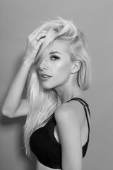 Fototapeta premium Black and white image. Sexy young blonde model in sexy black bra, holding a hand in her hair over gray background.