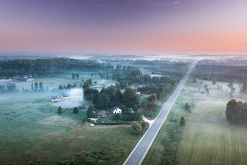 Highway going through meadow in mist surrounded by forest. Aerial view over picturesque landscape in fog at sunrise. 
