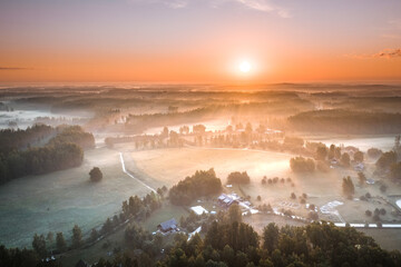 Highway going through meadow in mist surrounded by forest. Aerial view over picturesque landscape in fog at sunrise. 

