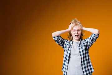 The young blonde girl in glasses in horror ruffles her hair from shock and unpleasant circumstances on an orange background in the studio