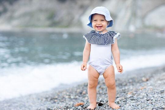 Cute Baby Girl Standing On The Sea Coast In A White Swimsuit And Laughting