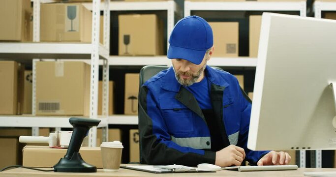 Caucasian postman in iniform and cap sitting at desk in postal office store and working at computer. Man typing on laptop. Portrait of male courier entering dta of invoice document in storage of boxes