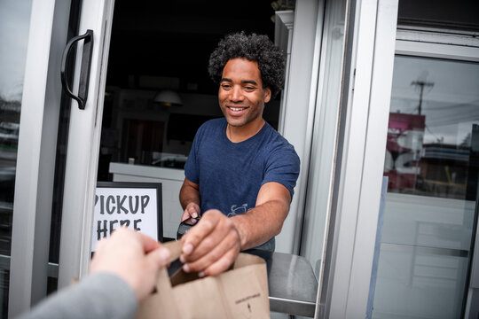 Male Business Owner Handing Takeout Food To Customer At Cafe Window