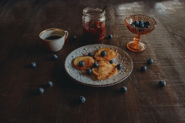 side view of a white vintage plate with pancakes and fresh blueberries and a cup of coffee with a jar of jam on a dark wooden table