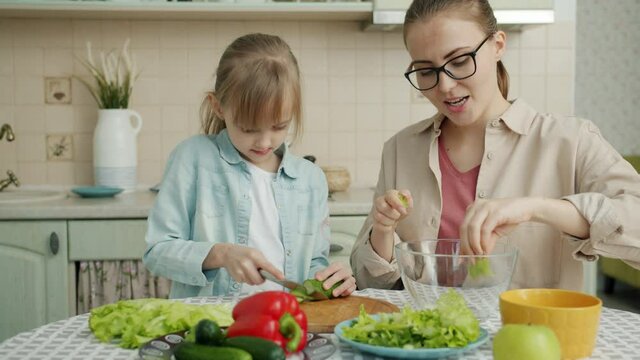 Adorable little girl is helping mother in kitchen cutting vegetables for salad and talking making dinner together. Healthy nutrition, lifestyle and people concept.