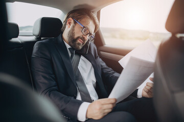 Successful businessman in formal suit sitting on backseat of car with working documents and talking...