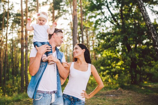 stylish young Family of mom, dad and daughter one year old blonde sitting near father on shoulders, outdoors outside the city in a park amid trees in summer. Wear jeans clothes. Family photo session