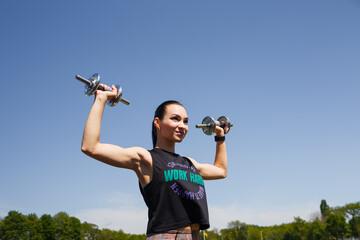 Fototapeta na wymiar Smiling girl standing on stadium green lawn with dumbbells in hands. Sport, physical training, healthy way of life, fitness concept.