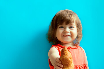 A little cheerful girl in pink dress with curly hair and blue eyes smiles, looks at the camera and holds a croissant in her outstretched hand. The child stands against the blue background.