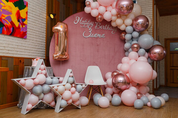 Beautiful festive decorations, pink and grey balloons arch, wooden stars, white chair and number one balloon on wooden round background. Little 1 year old girl birthday party photo zone.