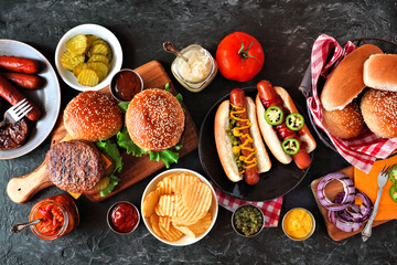 Summer BBQ food table with hot dog and hamburger buffet. Top down view over a dark slate background.