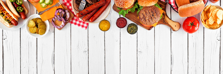 Summer BBQ food table scene with hot dog and hamburger buffet. Above view top border over a white...