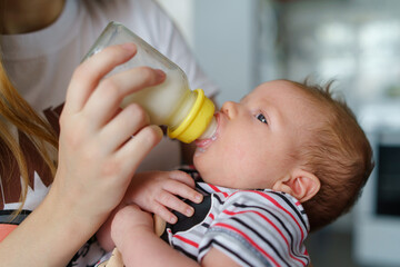 Mother holds her two moths old baby on the chair taking care at home - Caucasian woman with her newborn child bottle feeding milk on lap - Affectionate and bonding childhood and motherhood concept